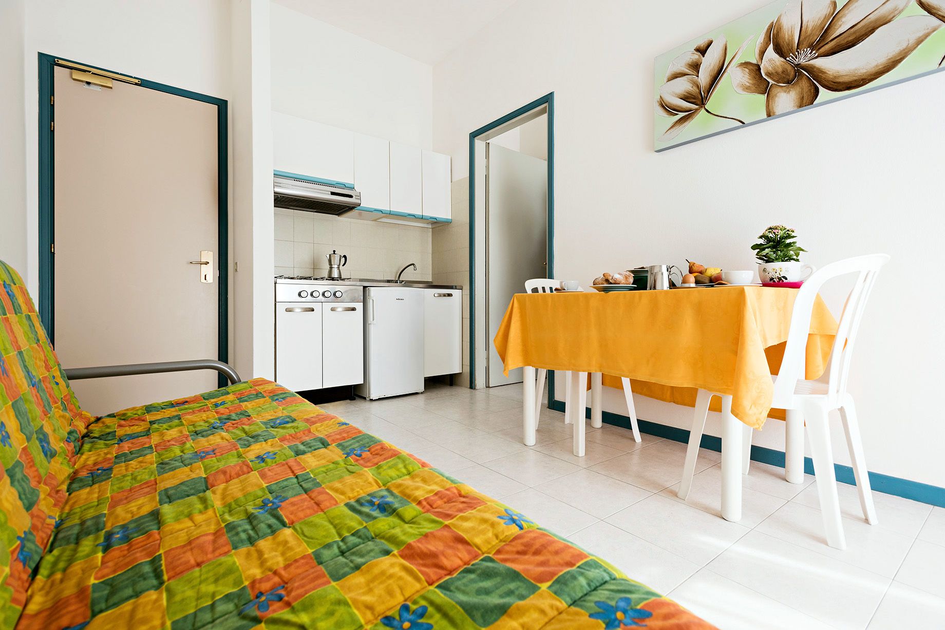 Een kamer met een bed, een eettafel met stoelen, een kitchenette en een schilderij aan de muur.