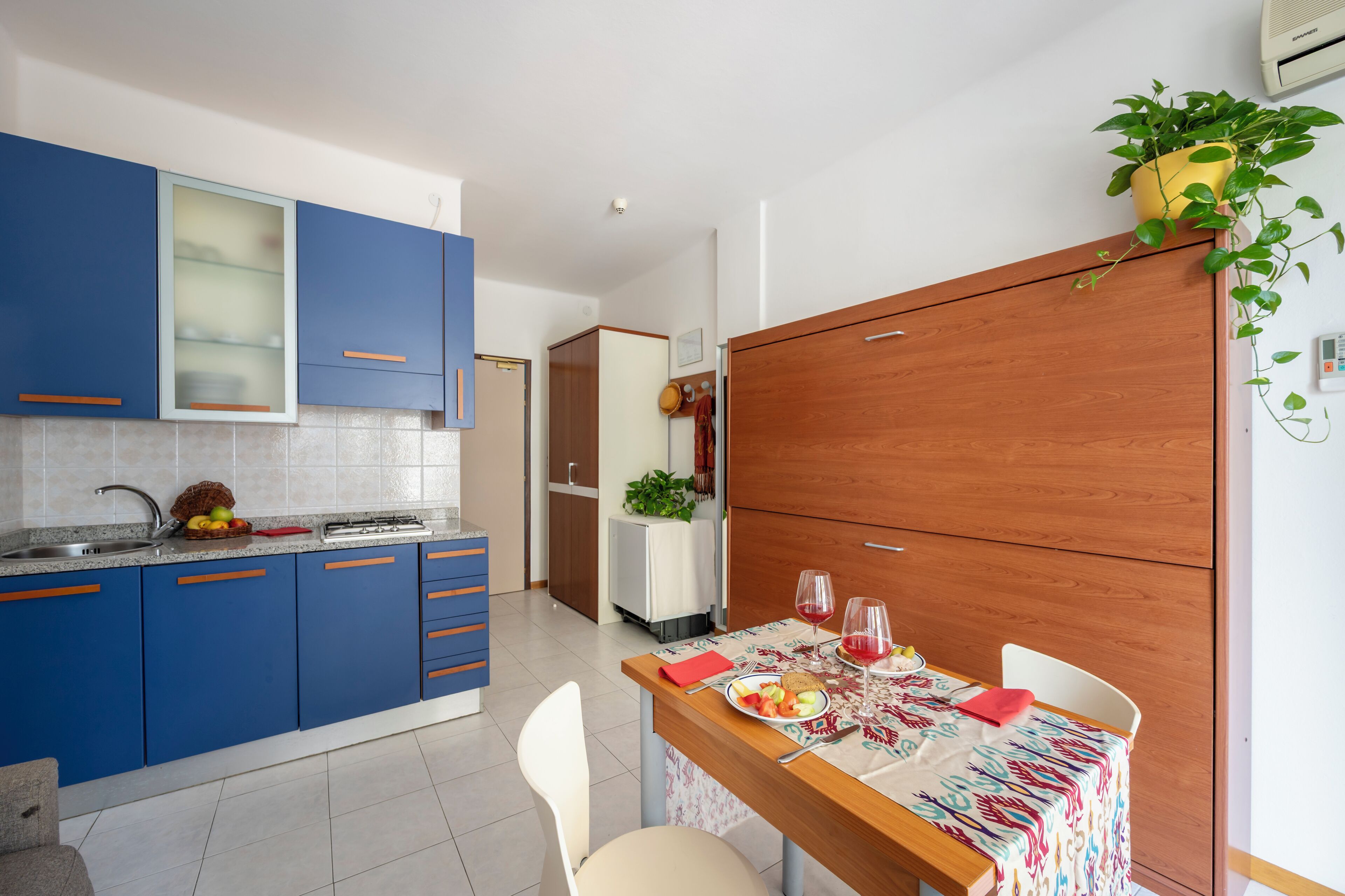 A modern kitchen with blue cabinets, a wooden dining table with a colorful tablecloth, and a potted plant on the wall.
