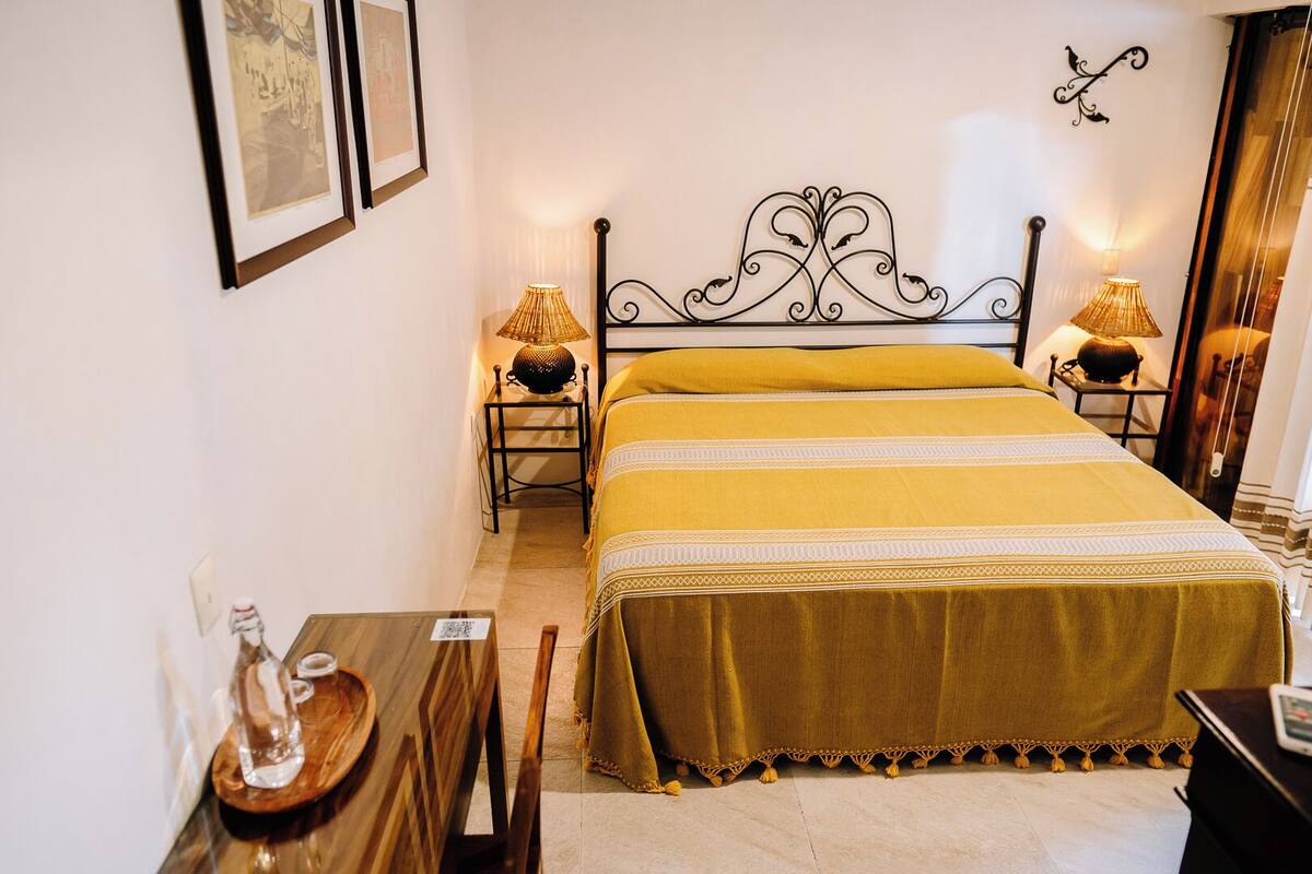 Un dormitorio ordenado con una cama, dos mesitas de noche con lámparas, una mesita auxiliar de madera y una silla.
