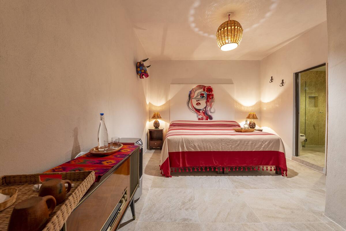 Un dormitorio con cama, lámparas de noche, un adorno en la pared, una mesita de mimbre y una puerta de cristal que da al baño.