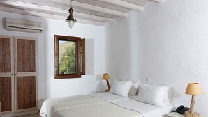 A bedroom with a bed, two bedside tables with lamps, a window with a view of greenery, and a wooden door. - Albatros Club Mykonos (Mykonos)
