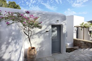 A white building with a grey door, a stone wall, and a potted plant with pink flowers. - Albatros Club Mykonos (Mykonos)