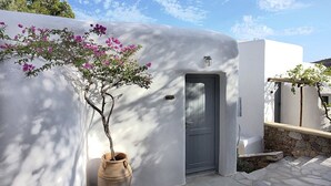 A white building with a grey door, a stone wall, and a potted plant with pink flowers. - Albatros Club Mykonos (Mykonos)