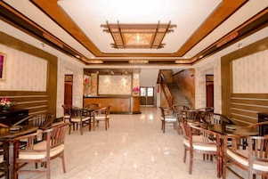 A hotel lobby with a reception desk, wooden chairs, a staircase, and a tiled floor.