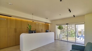 Uma área de recepção com um balcão branco, armários de madeira e uma grande porta de vidro que dá para uma área externa com plantas.