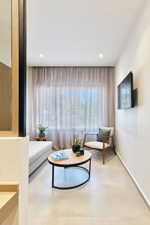A modern living room with a sofa, a round coffee table, a chair, a TV mounted on the wall, and a large window with sheer curtains. - Inea Studio (Chania)