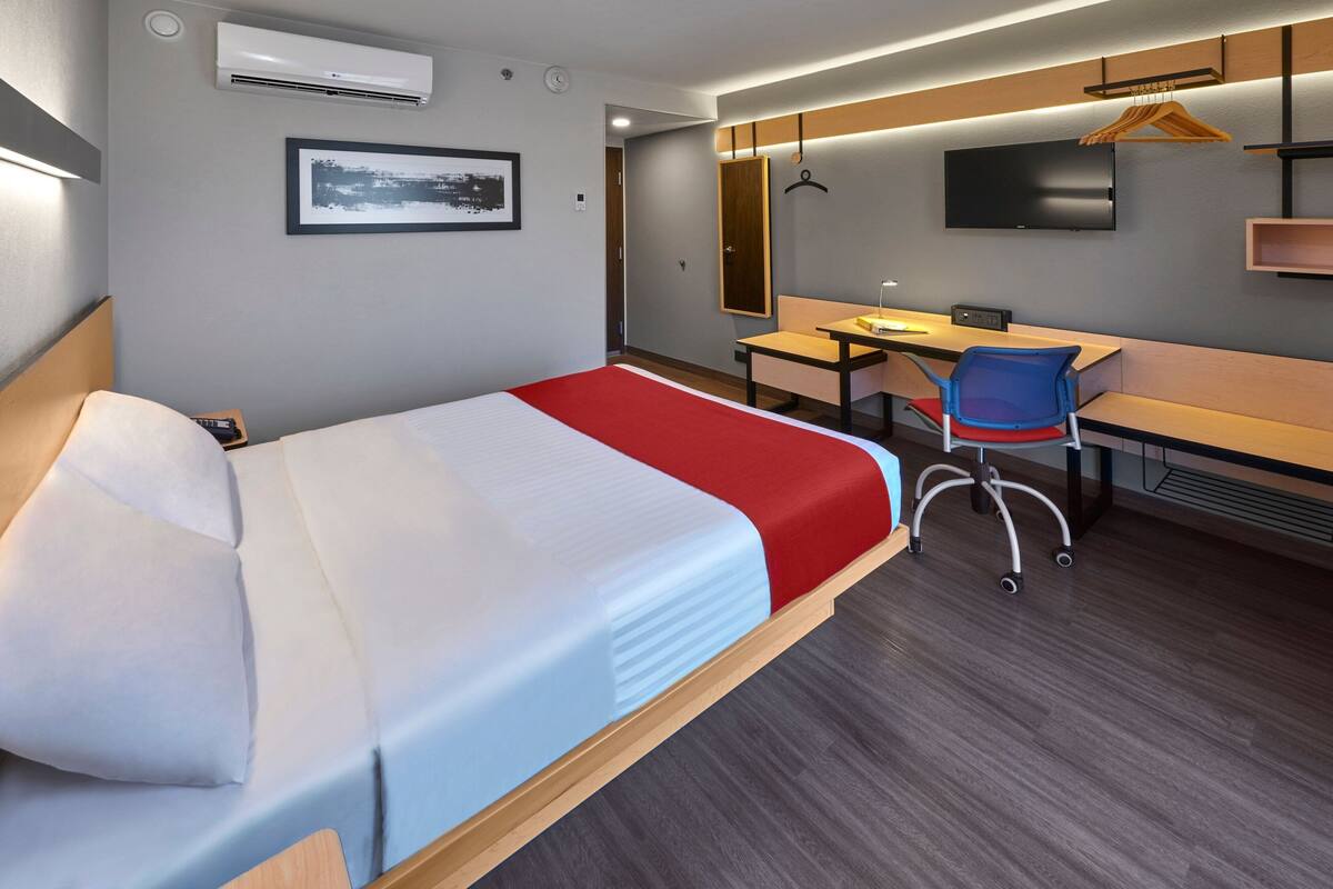 A hotel room with a bed, desk, chair, TV, and air conditioning unit.