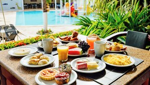 Un bufé de desayuno con diversos platos y bebidas, una piscina al fondo y asientos al aire libre.