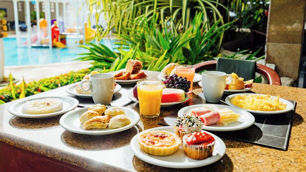 Un bufé de desayuno con diversos platos y bebidas, una piscina al fondo y plantas tropicales.