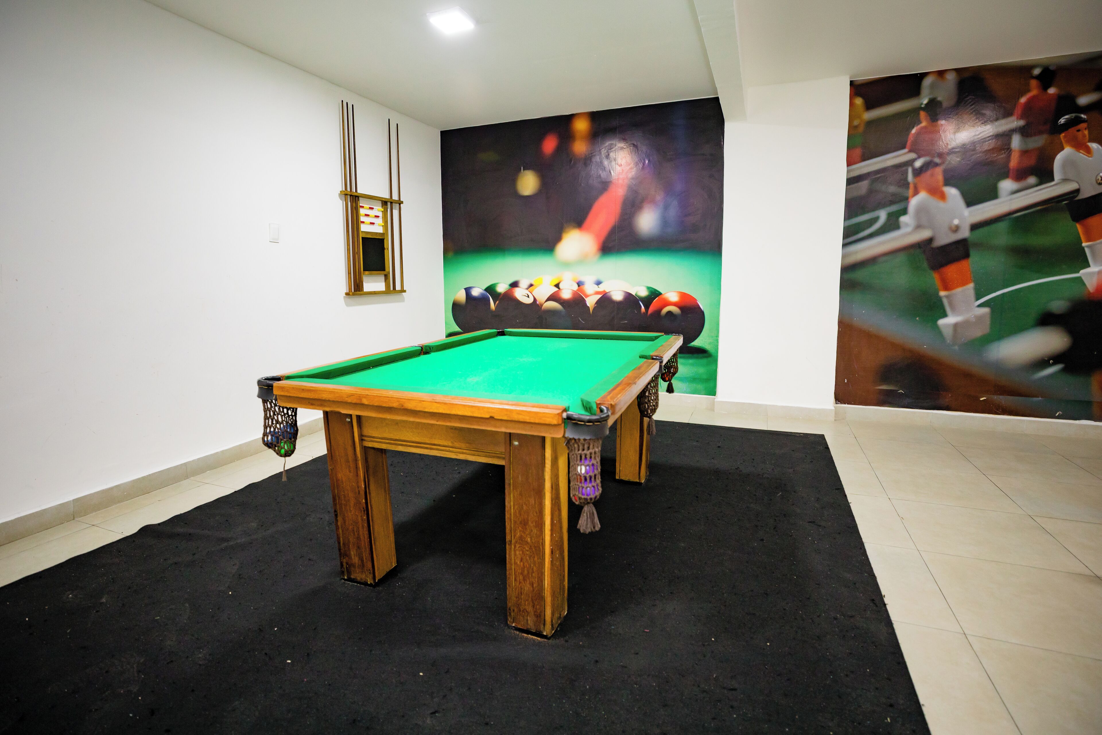 Una mesa de billar en una habitación con suelo de baldosas, un estante fijado a la pared y un gran mural con temática deportiva.