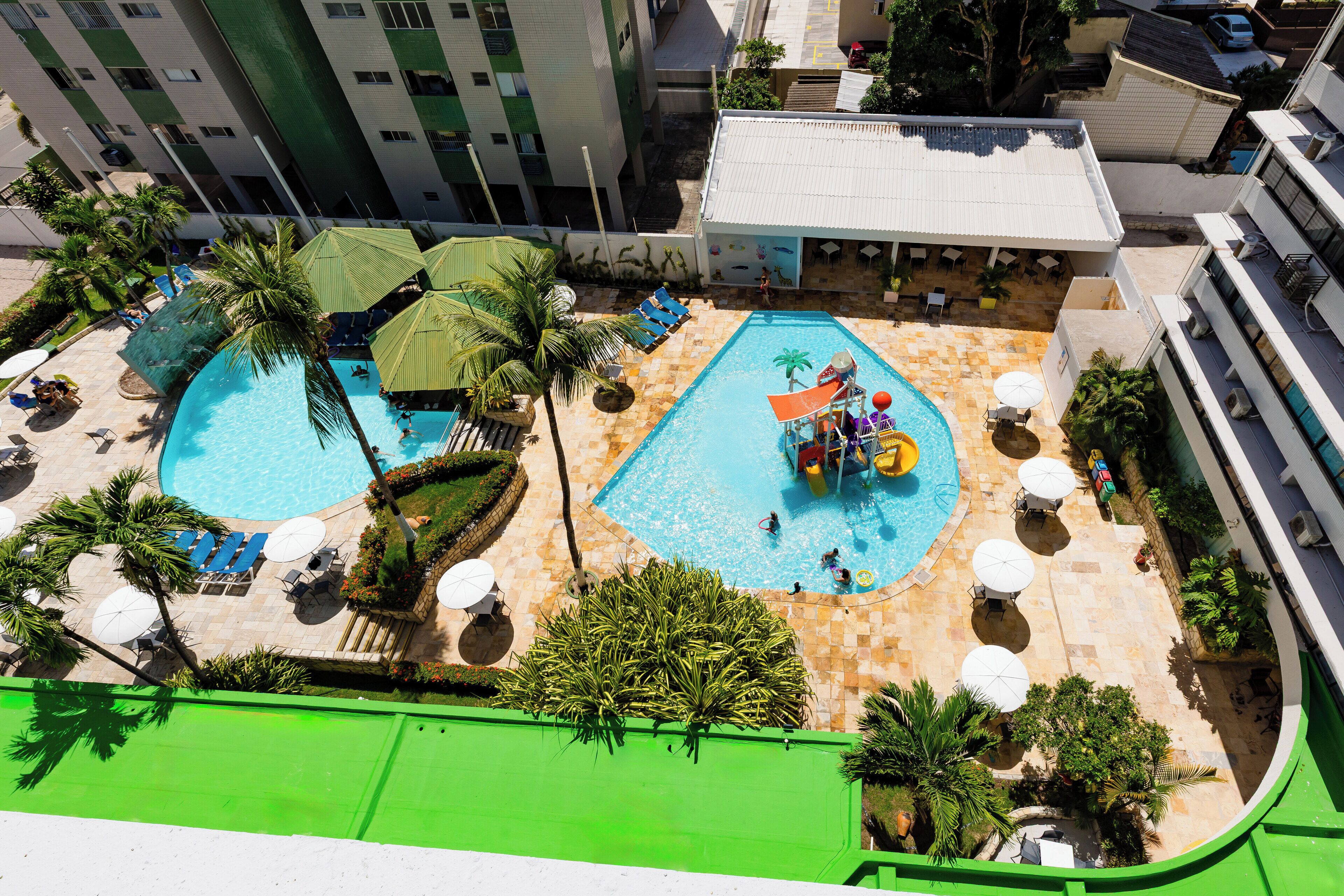 A rooftop pool area with two swimming pools, lounge chairs, umbrellas, and palm trees.