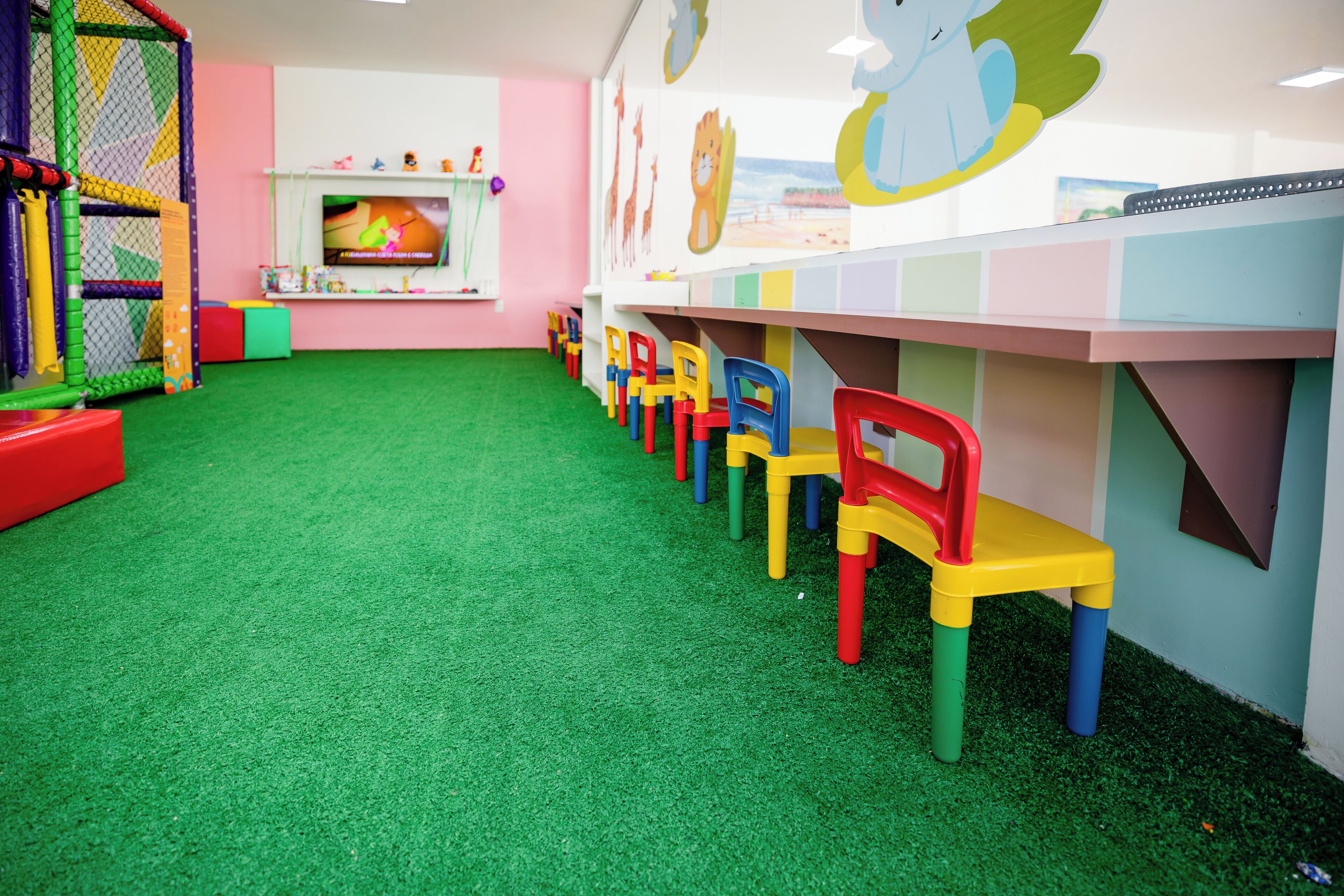 Un'area giochi per bambini con pavimento in erba sintetica verde, sedie di plastica colorate e una struttura giochi con una rete.