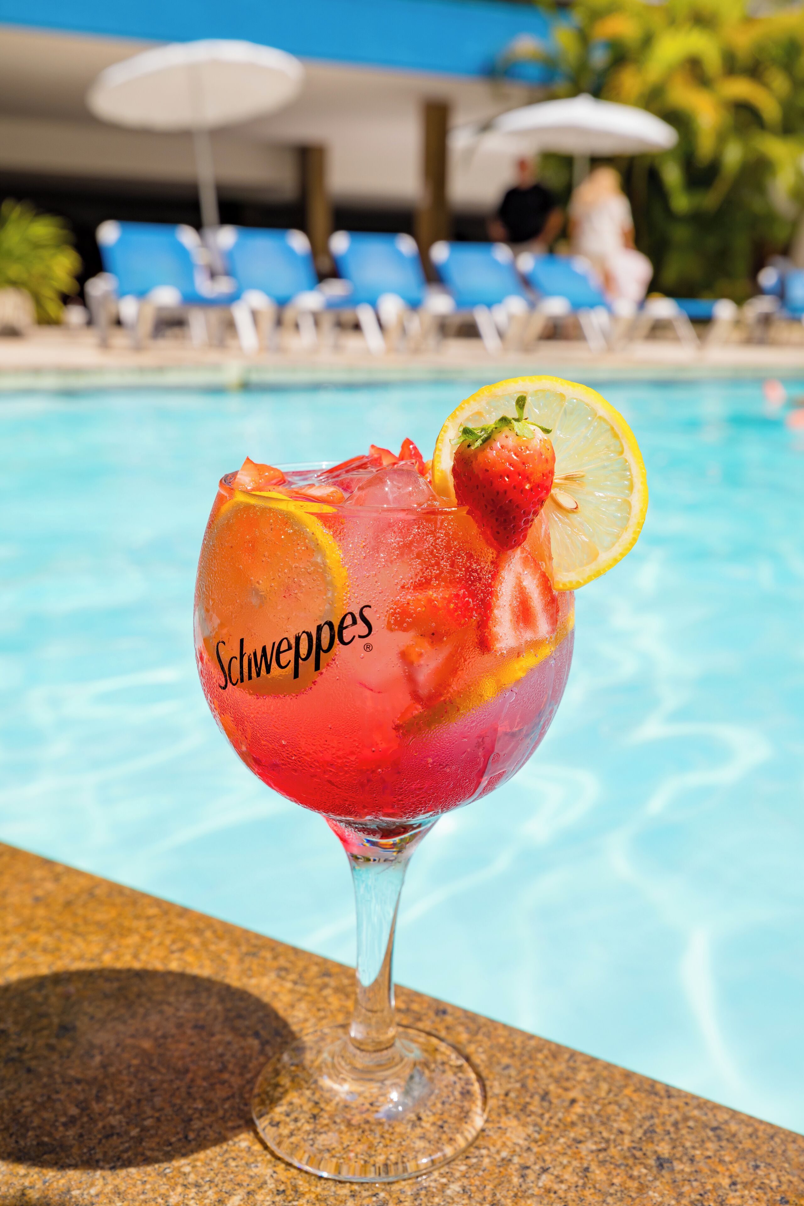 A cocktail glass with a strawberry and lemon slice garnish, labeled "Schweppes," on a poolside with blue lounge chairs and umbrellas in the background.