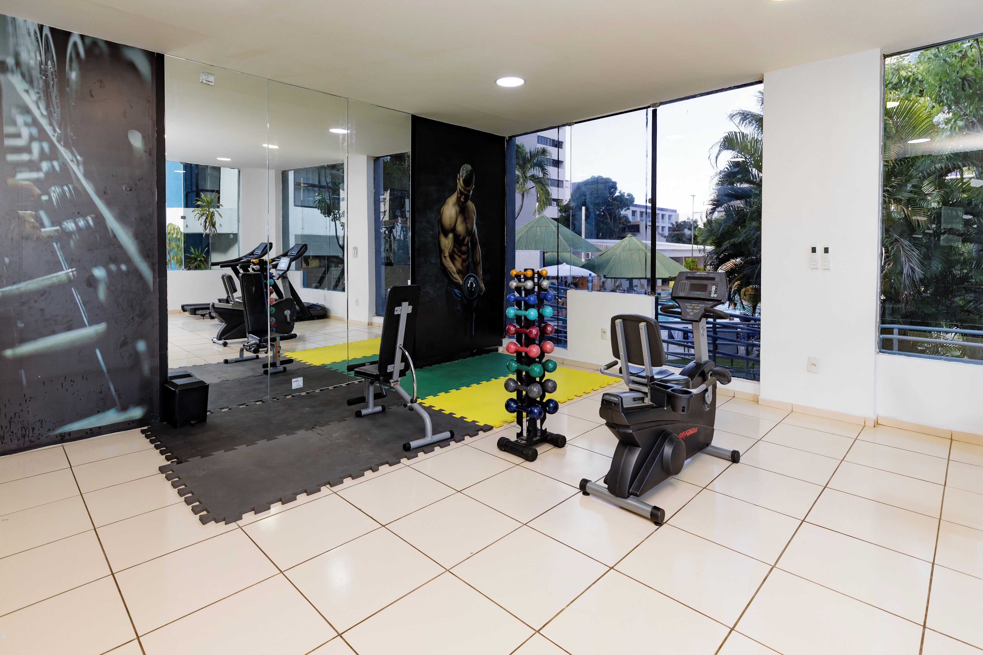 A gym with exercise equipment, a mirror wall, and large windows with a view of palm trees.