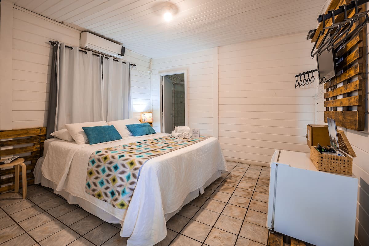 Quarto com cama, mesa de cabeceira, abajur e televisão presa na parede.