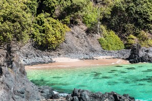A coastal landscape with rocky cliffs, clear turquoise waters, and a sandy beach. - Ecocharme Pousada Arquipelago (Fernando de Noronha)