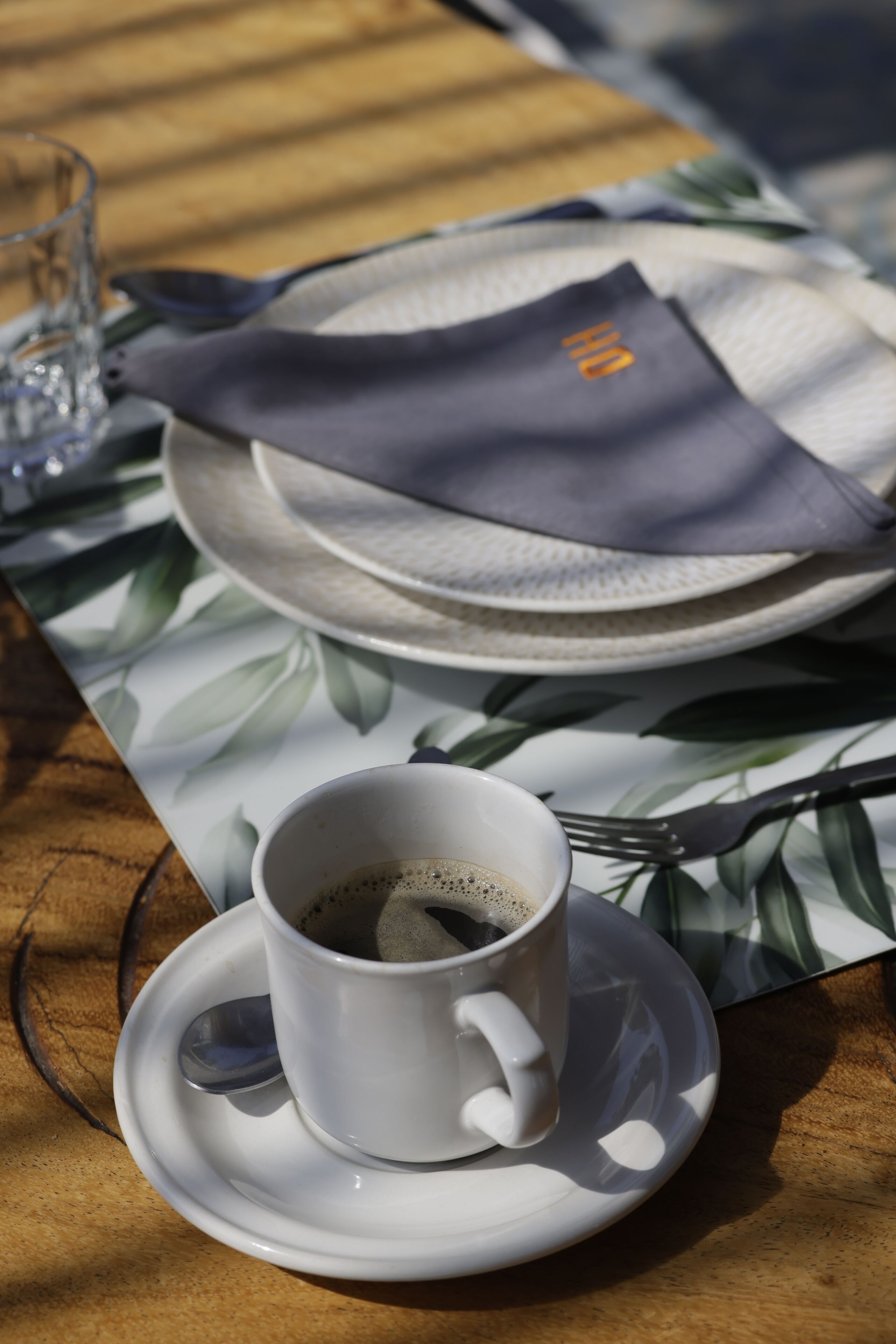 Una mesa con una taza de café blanco, un vaso de agua y una servilleta doblada sobre un mantel con estampado de hojas.