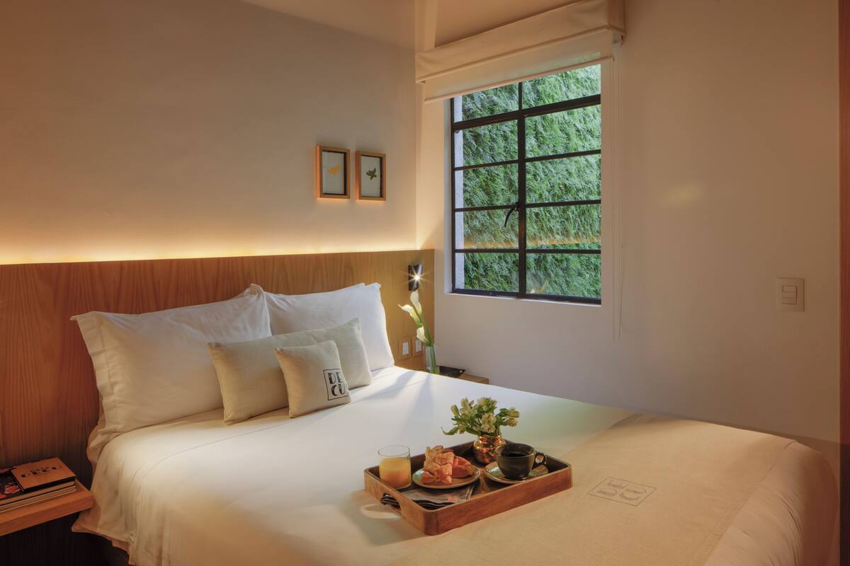 A neatly made bed with a tray of breakfast items, a window with a view of greenery, and a wooden headboard.