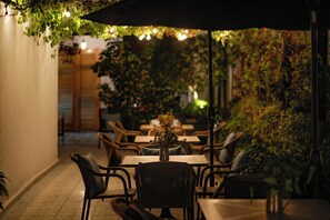 Un espacio al aire libre con mesas y sillas, rodeado de plantas y un ambiente cálido y tenue.