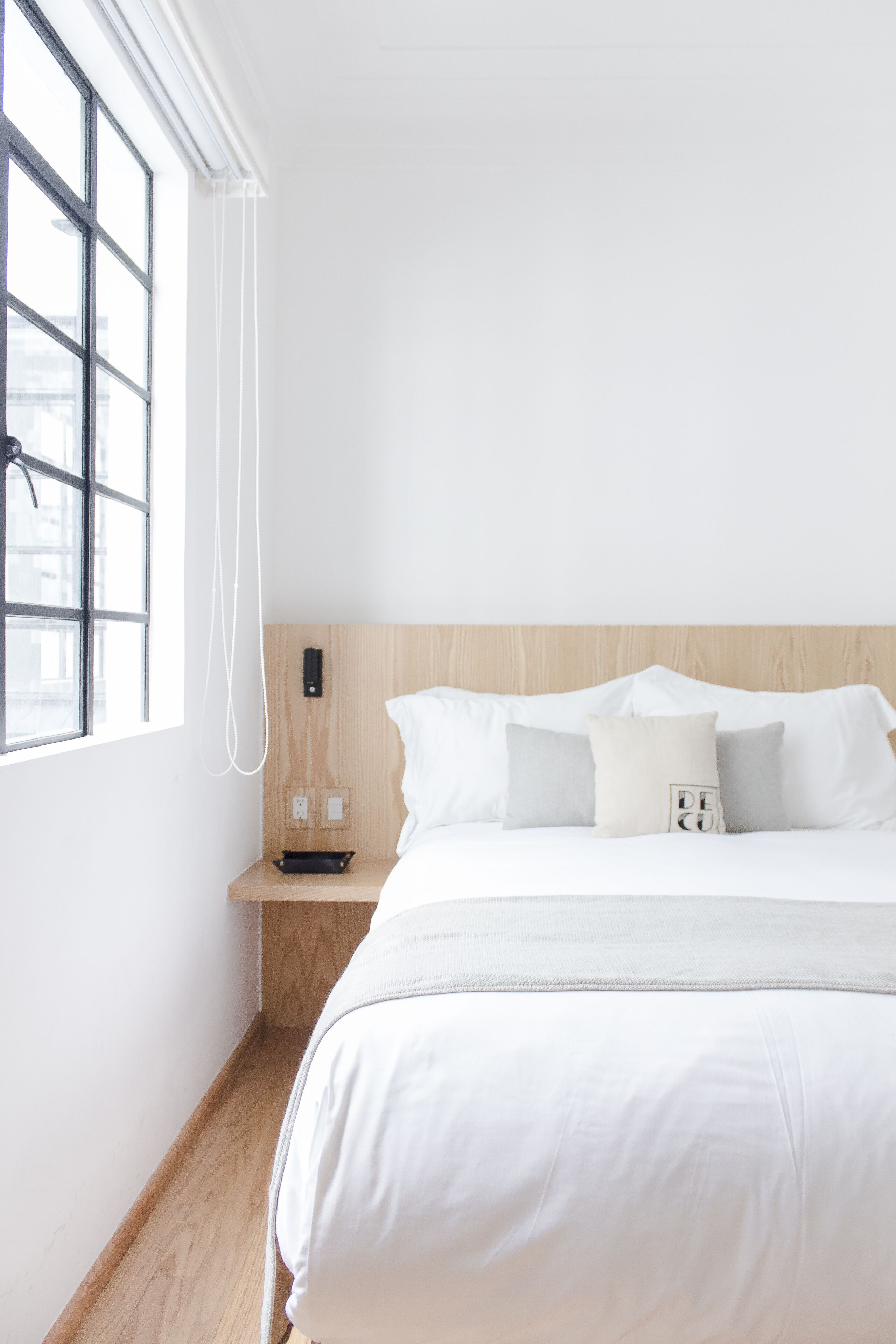 Une chambre d’hôtel au style minimaliste, dotée d’une grande fenêtre, d’une tête de lit en bois et d’un lit parfaitement fait avec du linge de lit blanc.