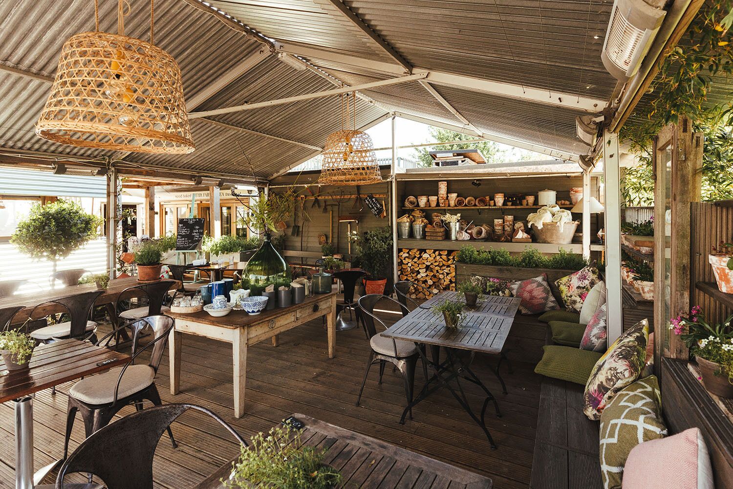 An outdoor café with wooden tables and chairs, hanging wicker lamps, and shelves with potted plants and pottery.