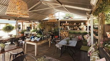 An outdoor cafรฉ with wooden tables and chairs, hanging wicker lamps, and shelves with potted plants and pottery.