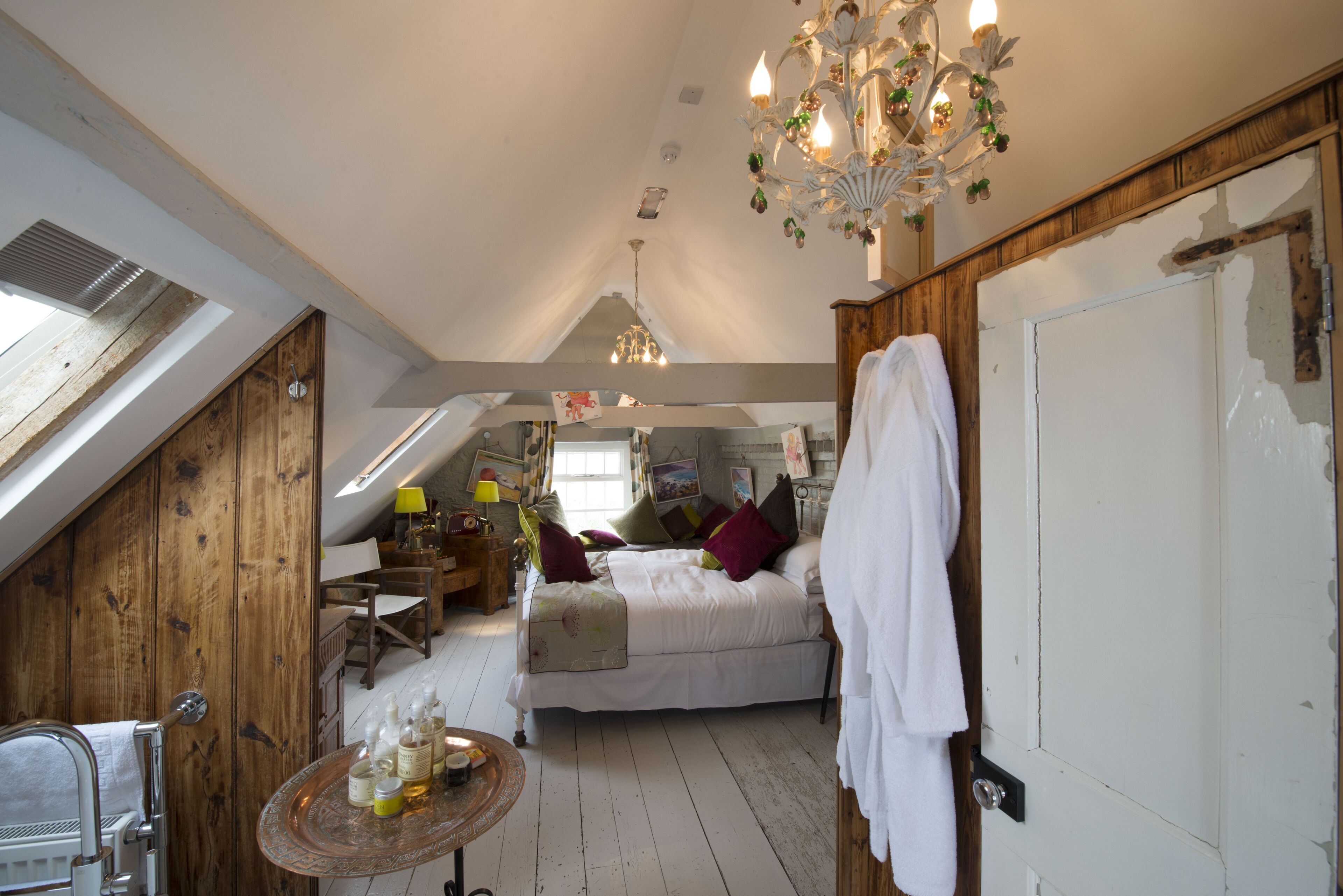 Ein Schlafzimmer mit schrägen Wänden, einem Bett mit Kissen, einer Kristallleuchtreiter, einer Holztür und einem kleinen Tisch mit Flaschen.