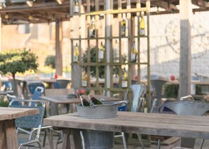 Un espacio de estar al aire libre con sillas y mesas de metal, un cubo de metal con botellas y una estructura de pérgola de madera al fondo.