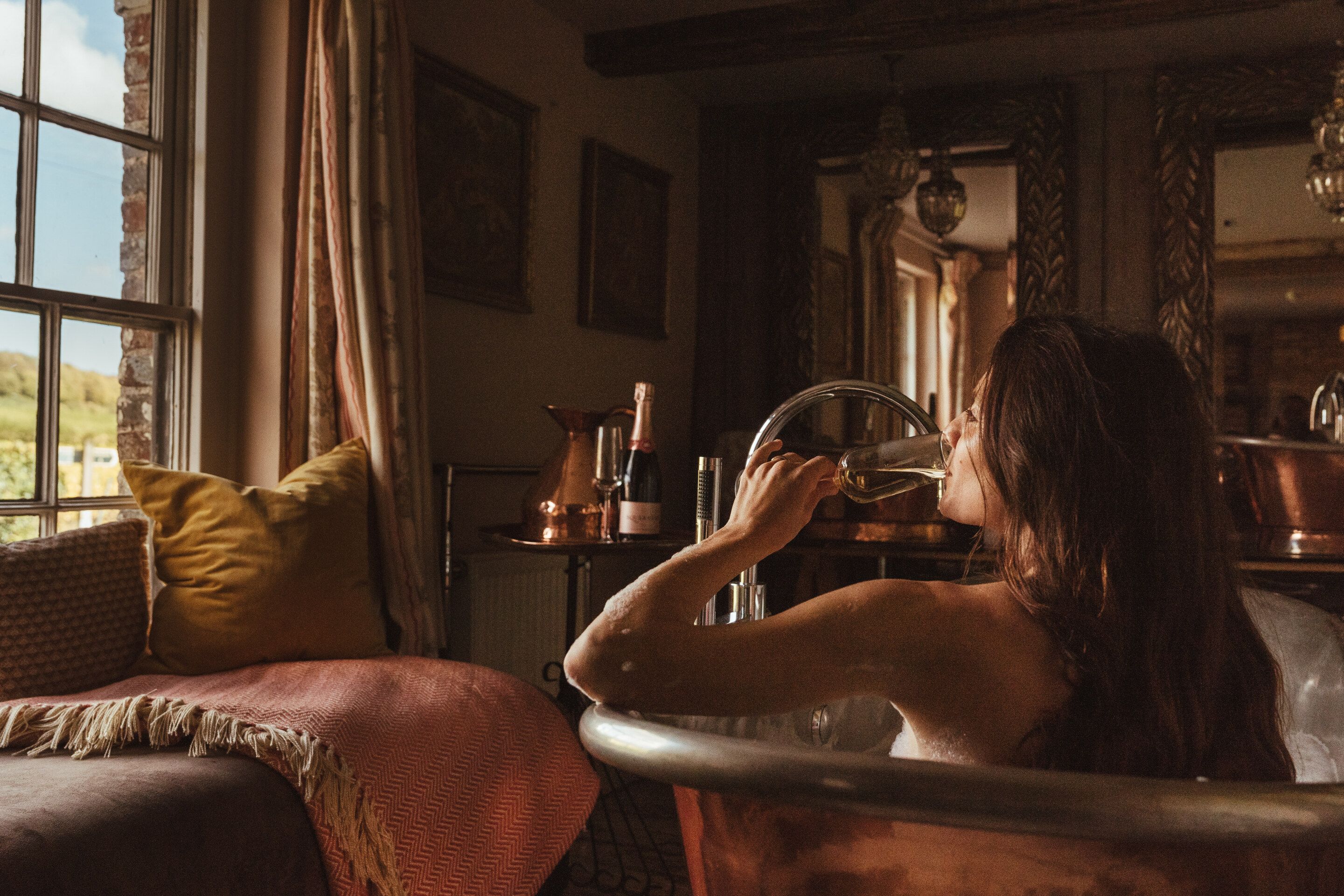 A person sitting by a window with curtains, holding a glass.
