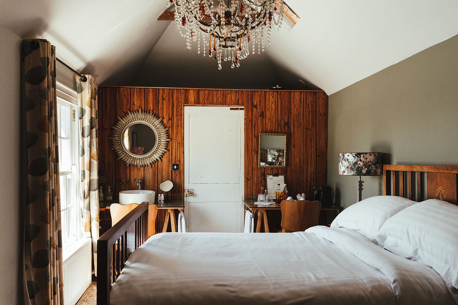 A bedroom with a wooden ceiling, a chandelier, a bed, a desk, a mirror, and a window with curtains.
