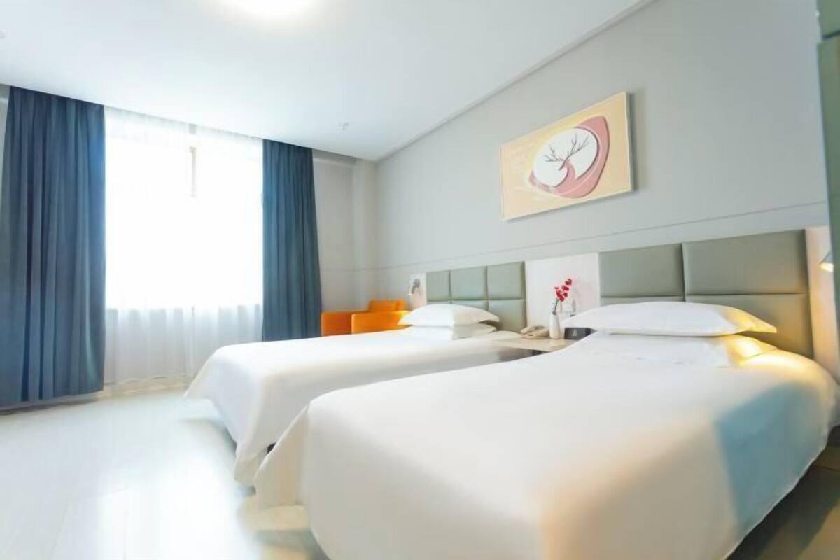 A hotel room with two beds, a large window with curtains, a painting on the wall, and a nightstand with a lamp.