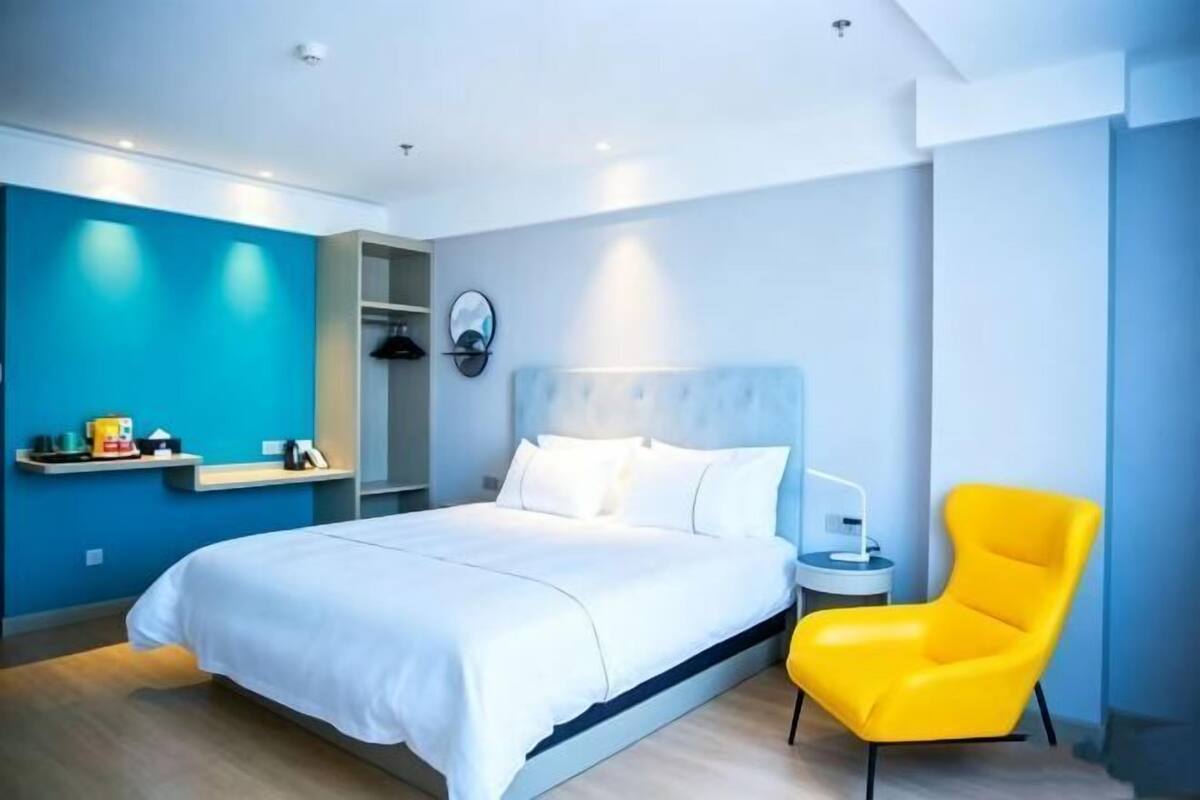 A modern hotel room with a bed, a bright yellow chair, a bedside table, and a wall-mounted shelf.