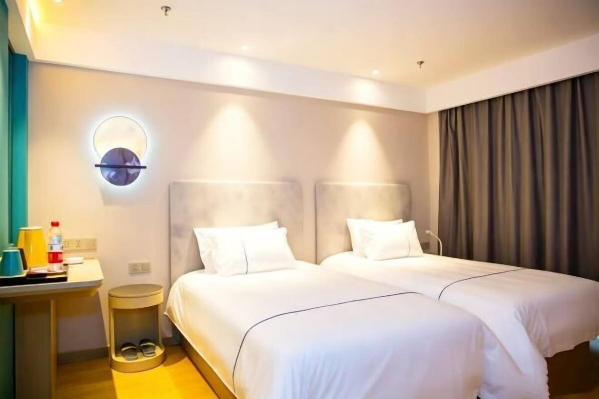 A hotel room with two beds, a bedside table, a round wall clock, a small stool with shoes, and a curtain.