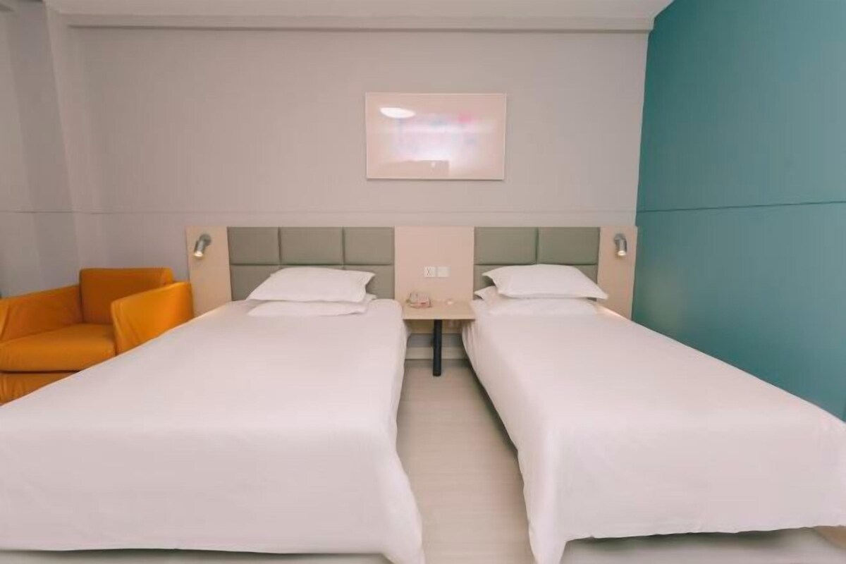 Une chambre d’hôtel avec deux lits simples, une petite table, une chaise et un luminaire fixé au mur.