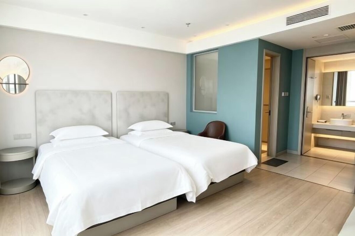 A modern hotel room with two beds, a bathroom with a large mirror, and a light-colored wooden floor.