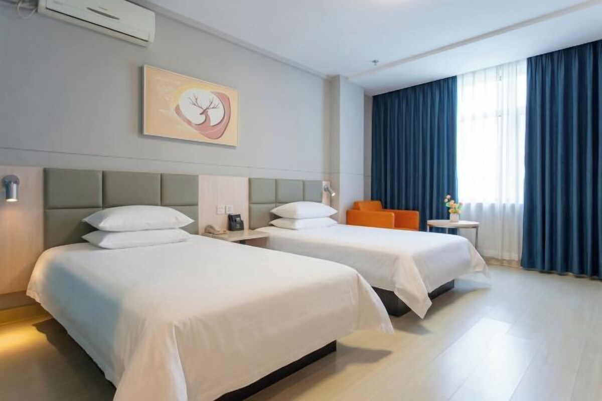 A hotel room with two beds, a headboard, a wall-mounted artwork, a window with curtains, and a small table with a vase.