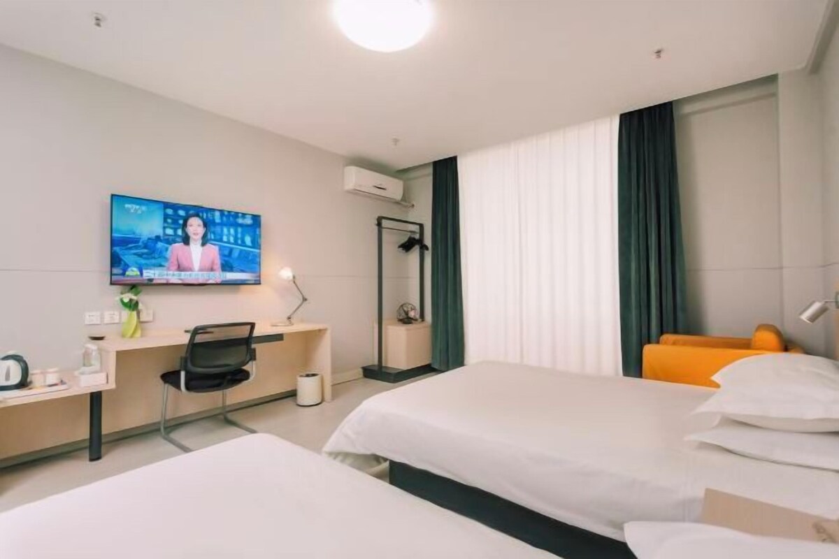 A hotel room with two beds, a desk with a chair, a TV mounted on the wall, and a window with curtains.