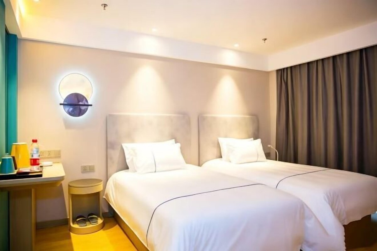 Une chambre d’hôtel avec deux lits, une table de chevet, une horloge ronde fixée au mur, un petit tabouret avec des chaussures et un rideau.