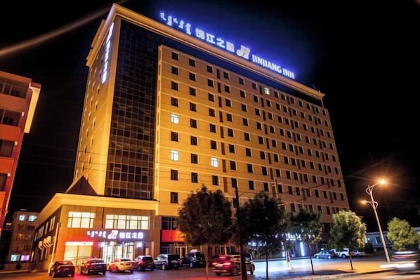 A multi-story hotel with a prominent sign at the top, a ground-level entrance, and a parking area with cars.
