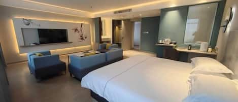 A modern hotel room with a large bed, a flat-screen TV mounted on the wall, a seating area with sofas, and a small table with a lamp.