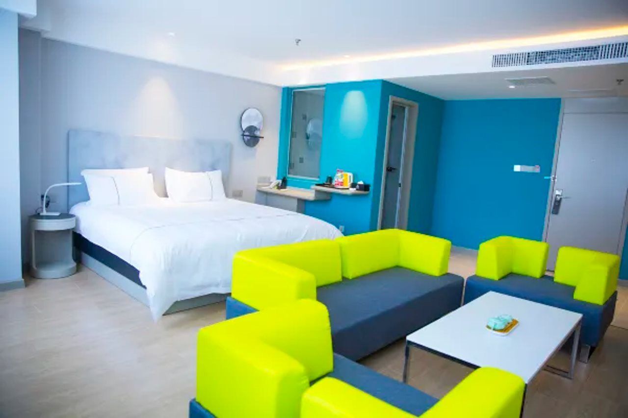 Una camera d'hotel moderna con un letto grande, un'area salotto con divani giallo e blu vivaci, un tavolino e la porta del bagno.