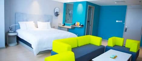 Una habitación de hotel moderna con una cama grande, un área de descanso con sofás azul y amarillo brillantes, una mesita y la puerta del baño.