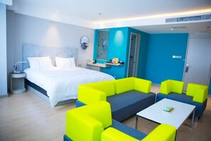 Een moderne hotelkamer met een groot bed, een zithoek met heldergele en blauwe banken, een tafeltje en een badkamers deur.
