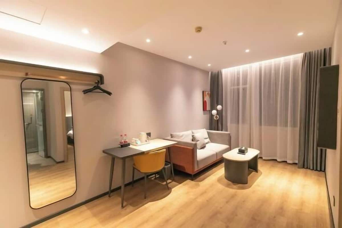 A modern hotel room with a sofa, a small table, a desk with a chair, a mirror, and a TV.
