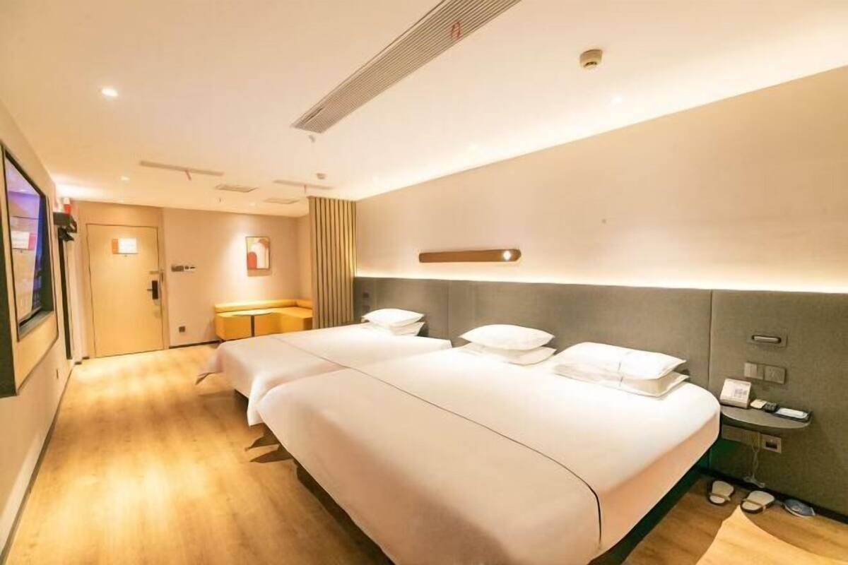 A hotel room with two beds, a bedside table, a small desk, and a wall-mounted light fixture.