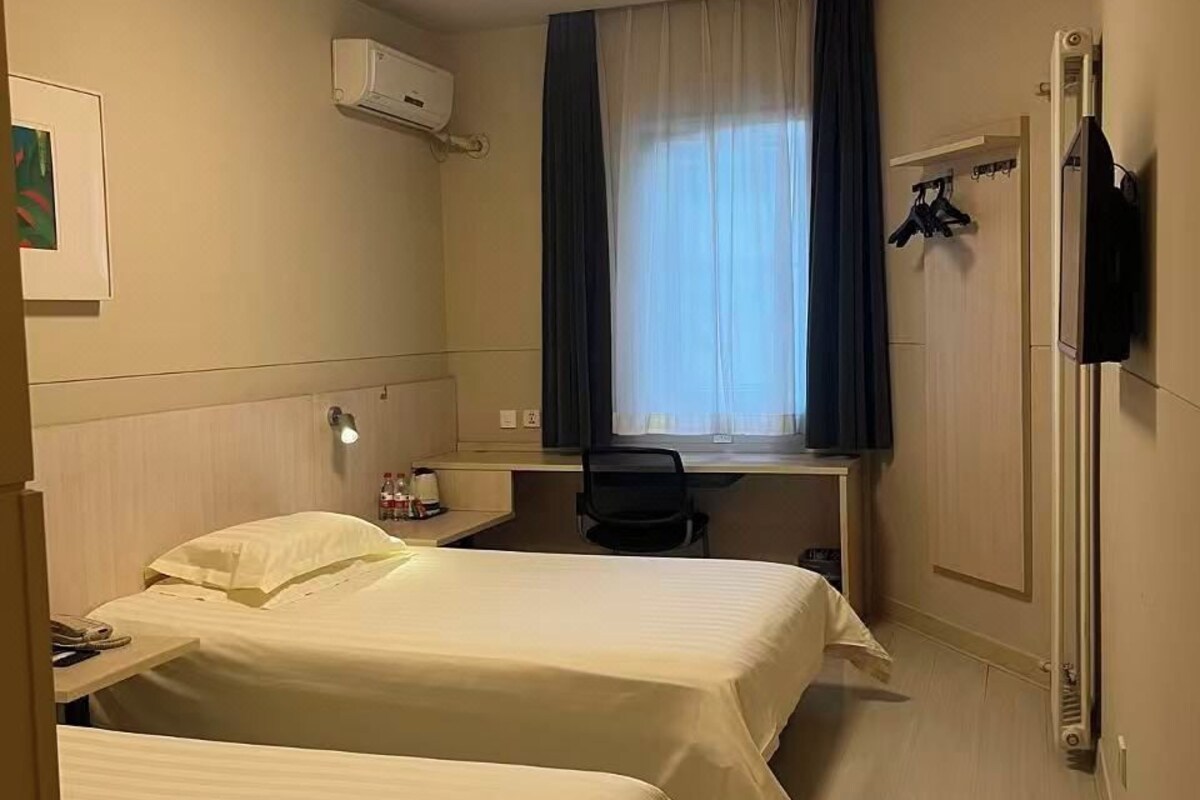 Habitación de hotel con dos camas, un escritorio, una silla, una ventana con cortinas, un equipo de aire acondicionado y un perchero.