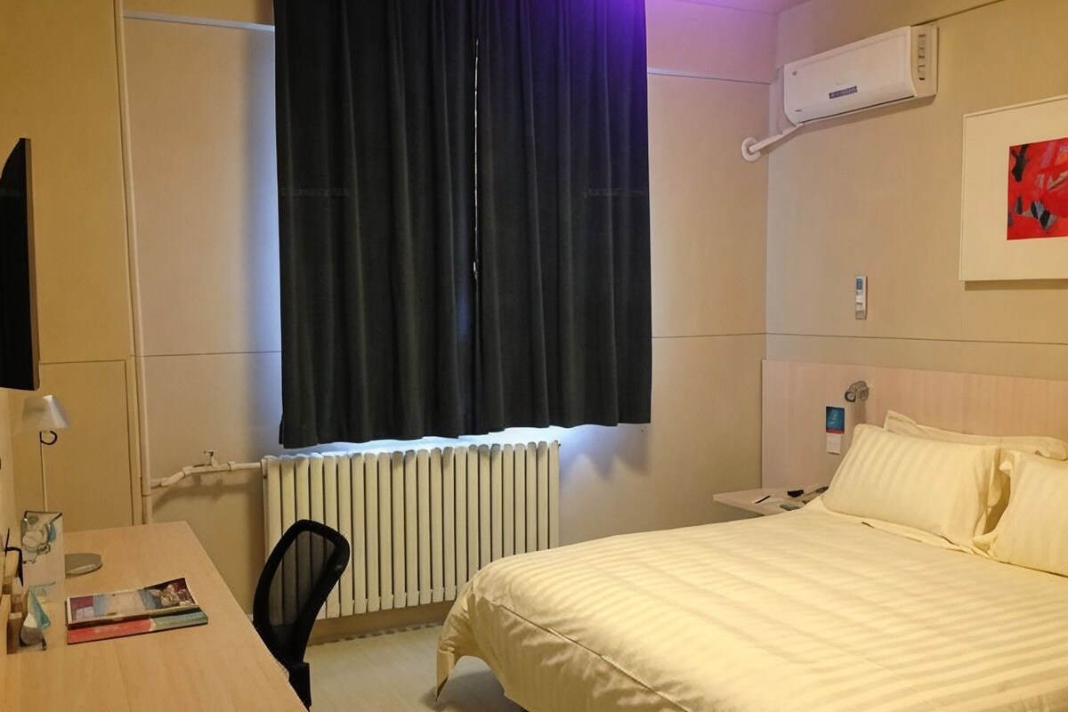 Habitación de hotel con cama, escritorio, silla, radiador y ventana con cortinas.