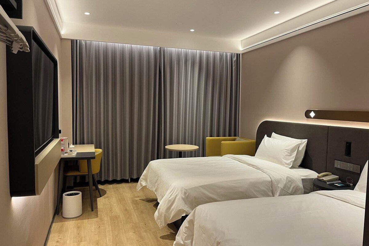 Habitación de hotel con dos camas, un escritorio, una silla, un televisor y un ventanal con cortinas.