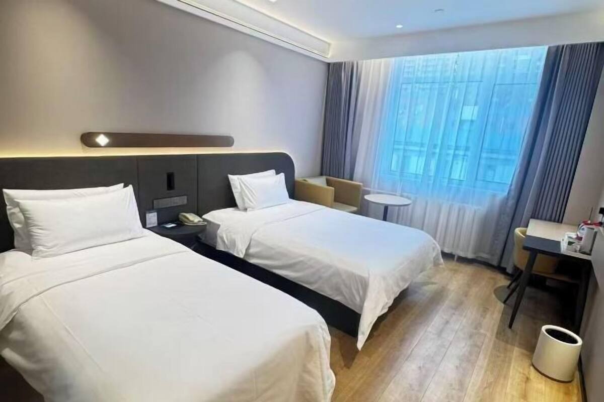Una habitación de hotel con dos camas, una mesita, una silla y una ventana con cortinas.