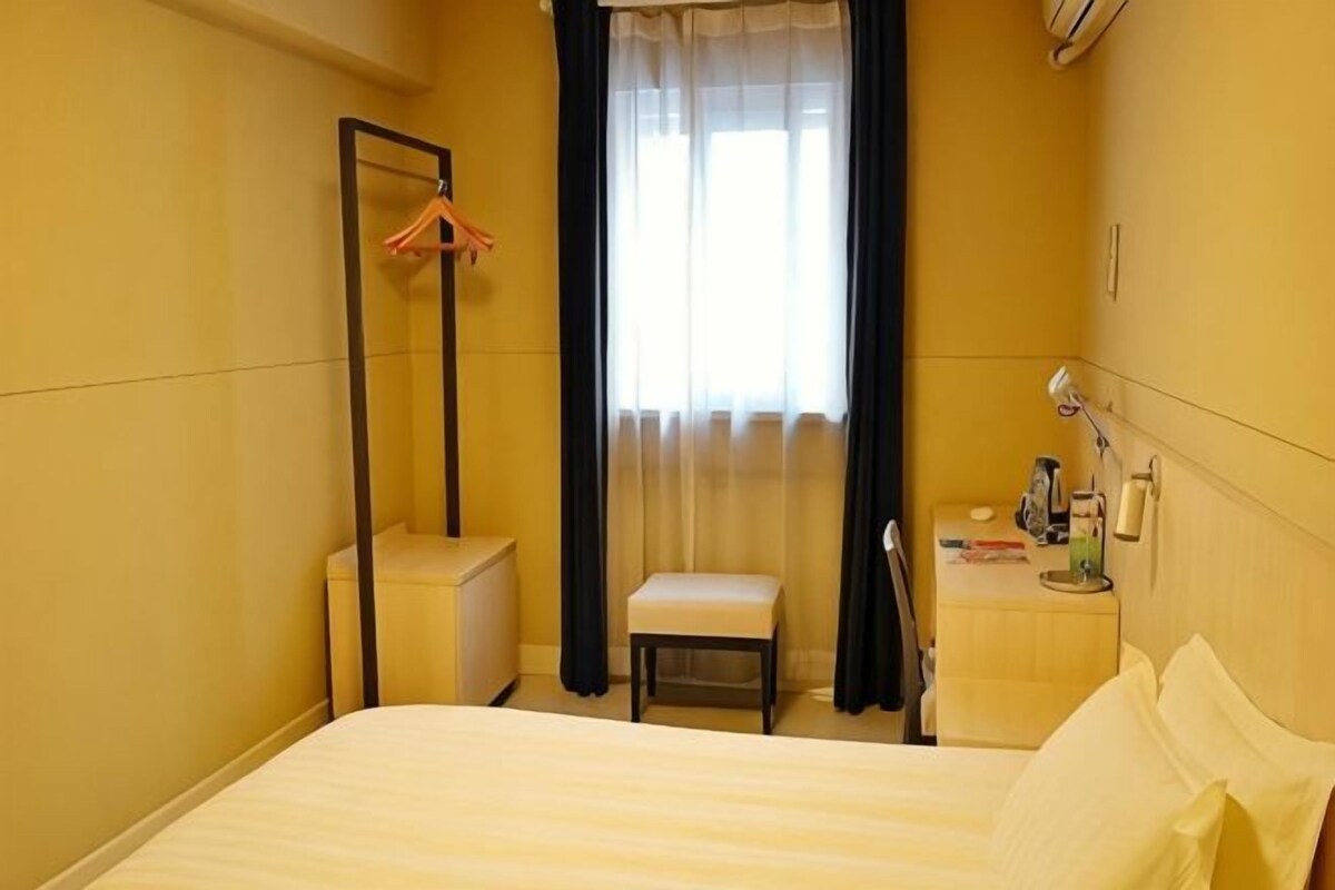 Ein Hotelzimmer mit Bett, Nachttisch mit Lampe, Stuhl, Kleiderhaken und einem Fenster mit Vorhängen.